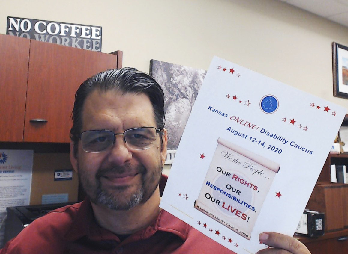 A white man with very dark hair, and a greying goatee, glasses, and a red top is smiling as he holds up a flyer. The flyer has red stars scattered around the white page. Flyer text: Kansas ONLINE! Disability Caucus August 12-14, 2020. Beneath the text is the image of the 2020 Caucus logo Logo: A scroll opened and showing text. Logo text: We the people. Our rights. Our responsibilities. Our lives! Background: A desk with a cabinet. On top of the cabinet is a sign that says "No Coffee No Workee"